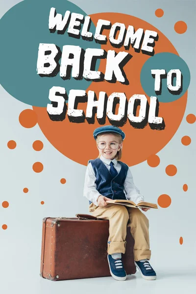 Lächelndes Kind in Retro-Weste und Mütze auf Koffer sitzend und Buch auf grauem Hintergrund mit Schulabbildung haltend — Stockfoto