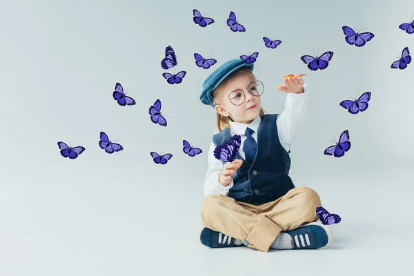 Entzückendes Kind sitzt mit überkreuzten Beinen auf dem Boden zwischen lila Feen-Schmetterlingen auf grauem Hintergrund — Stockfoto