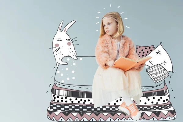 Verträumtes Kind in Kunstpelzmantel und Rock sitzt auf Fantasiebett mit Kissen neben Kaninchen und hält Buch isoliert auf grau — Stockfoto