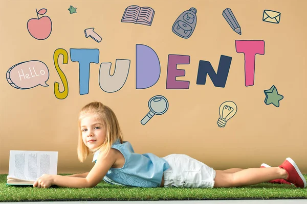 Cute kid lying on grass rug and holding book on beige background with student lettering — Stock Photo