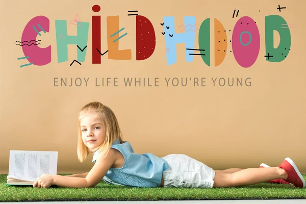 Мила дитина лежить на трав'яному килимку і тримає книгу на бежевому фоні з дитячою ілюстрацією — стокове фото