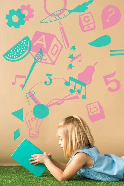 Smiling and cute child lying on grass rug and holding book on beige background with creative illustration — Stock Photo