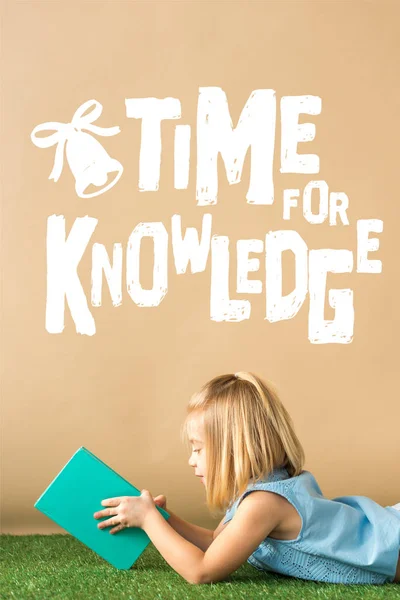 Smiling and cute child lying on grass rug and holding book on beige background with white time for knowledge lettering — Stock Photo