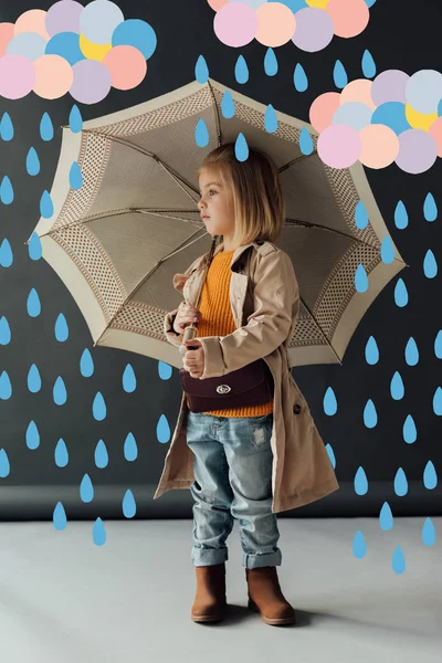 Ernstes Kind in Trenchcoat und Jeans mit Regenschirm im Märchenregen und wegschauendem Blick — Stockfoto