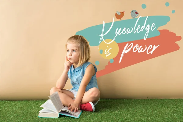 Thoughtful cute child sitting with crossed legs on grass rug and holding book on beige background with knowledge is power lettering — Stock Photo