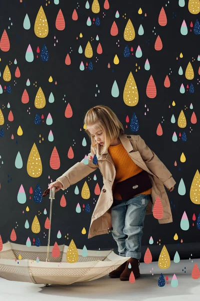 Серьезный ребенок в плаще и джинсах с зонтиком под волшебным дождем — стоковое фото