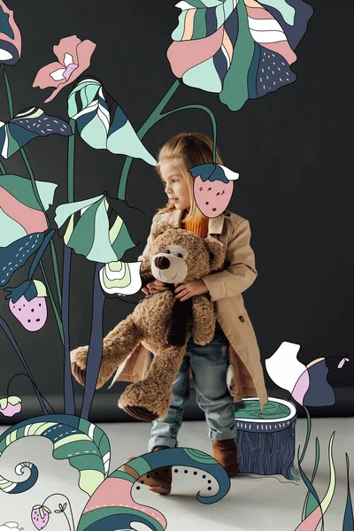 Cute child in trench coat and jeans holding teddy bear on black background with fairy plants illustration — Stock Photo
