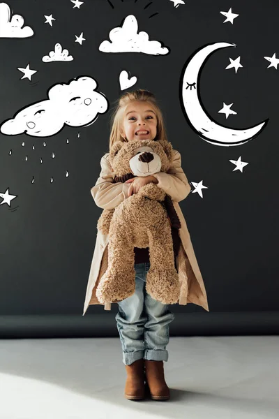 Happy and cute child in trench coat and jeans hugging teddy bear on black background with magic moon, stars and rainy cloud illustration — Stock Photo