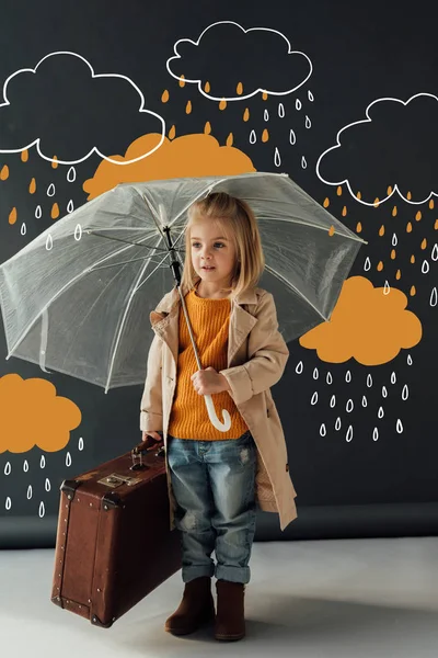 Child in trench coat and jeans holding umbrella and leather suitcase under fantasy rain on black background — Stock Photo