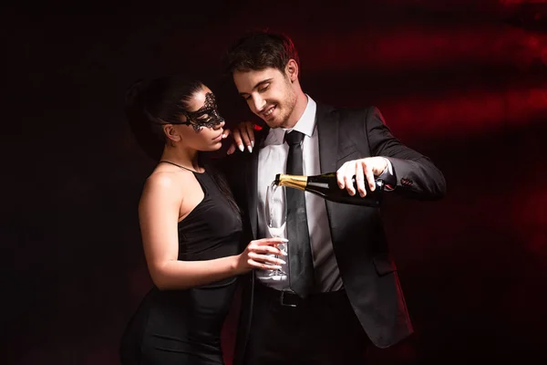 Homme souriant en tenue formelle versant champagne à la femme en robe et masque sur noir — Photo de stock