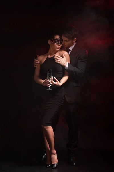 Visão de comprimento total do casal sensual bdsm com vidro em champanhe abraçando no preto — Fotografia de Stock