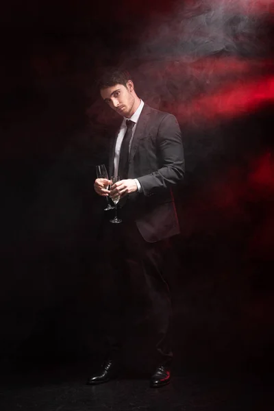 Full length view of handsome man in formal wear holding wineglasses in smoke on black — Stock Photo