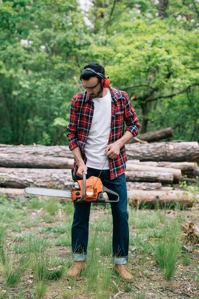 Lumberjack in checkered shirt and noise-canceling headphones adjusting chainsaw — Stock Photo