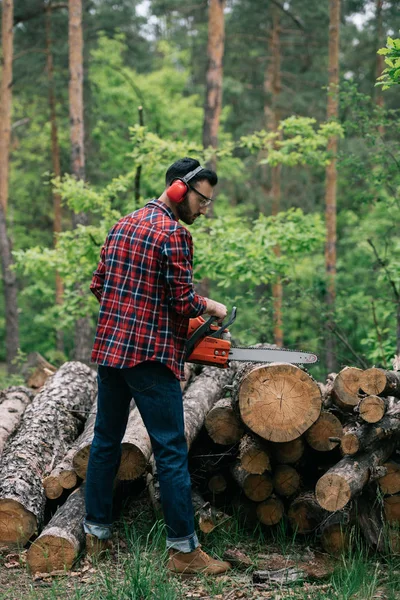 Bärtiger Holzfäller im Gehörschutz schneidet Stamm mit Kettensäge im Wald — Stockfoto