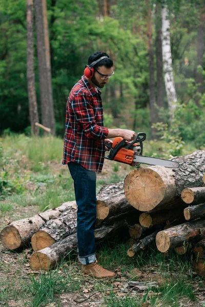 Bearded lumberjack in noise-canceling headphones cutting wood with chainsaw in forest — Stock Photo