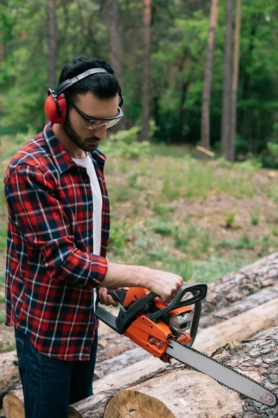 Aufmerksame Holzfäller im karierten Hemd schneiden mit Kettensäge Holz im Wald — Stockfoto