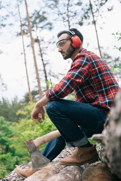 Nachdenklicher Holzfäller hält Axt in der Hand und schaut weg, während er im Wald auf Baumstämmen sitzt — Stockfoto