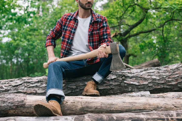 Partial view of lumberjack holding ax while sitting on logs in forest and holding ax — Stock Photo