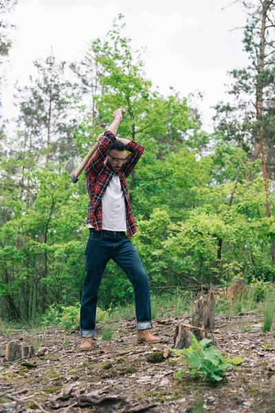 Bûcheron en chemise à carreaux et jeans en denim coupant le bois à la hache en forêt — Photo de stock