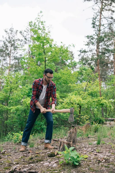 Lumberjack in checkered shirt and denim jeans cutting wood with ax in forest — Stock Photo