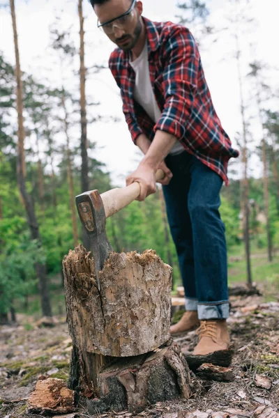 Selective focus of lumberman in plaid shirt and denim jeans cutting wood with ax in forest — Stock Photo