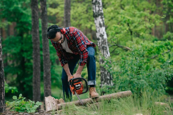 Taglialegna in camicia scozzese e jeans jeans taglio legno con motosega nel bosco — Foto stock