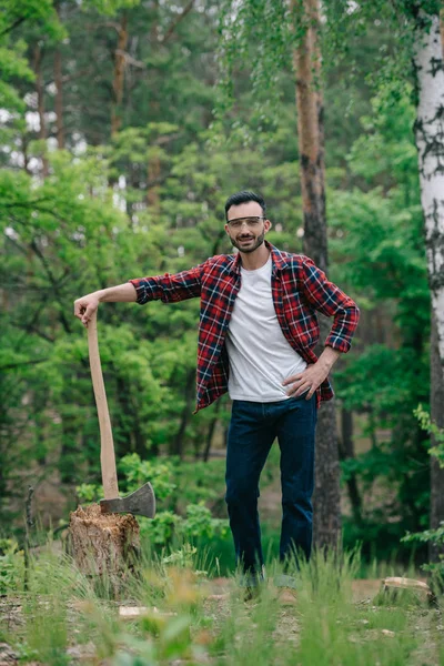 Leñador alegre en camisa a cuadros y vaqueros vaqueros sosteniendo hacha mientras está de pie con la mano en la cadera y sonriendo a la cámara - foto de stock