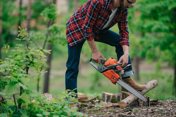 Vue partielle du bûcheron en chemise à carreaux et jean en denim coupant le bois avec tronçonneuse en forêt — Photo de stock