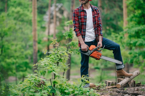 Partial view of lumberman in plaid shirt and denim jeans standing with chainsaw in forest — Stock Photo