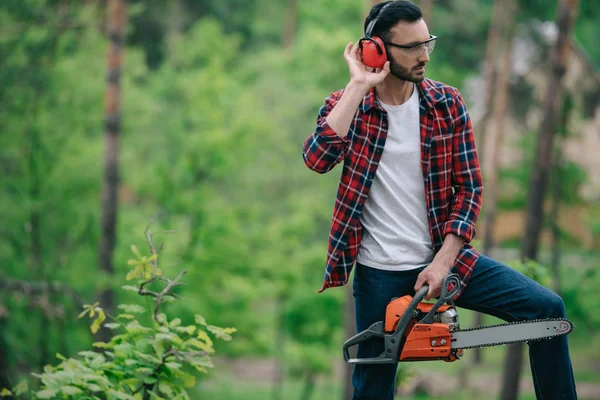 Bûcheron en chemise à carreaux debout avec tronçonneuse dans la forêt et touchant casque anti-bruit — Photo de stock