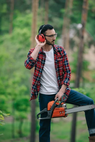 Lenhador grave em camisa xadrez em pé com motosserra na floresta e tocando protetores auditivos — Fotografia de Stock