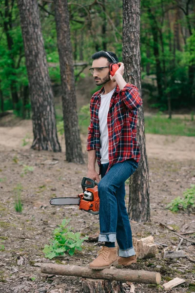 Thoughtful lumberjack holding chainsaw, touching noise-canceling headphones and looking away — Stock Photo