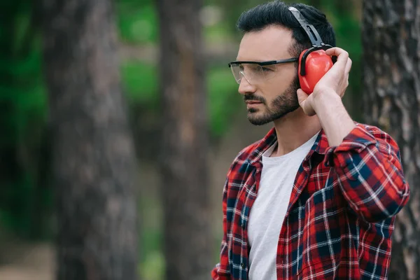 Pensive lumberjack touching noise-canceling headphones and looking away in forest — Stock Photo