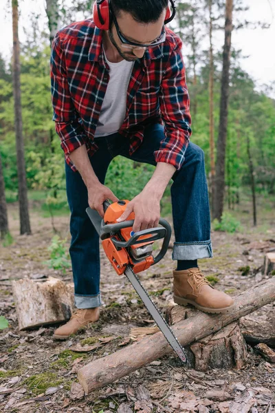 Leñador en camisa a cuadros y vaqueros tejanos cortando madera con motosierra en el bosque - foto de stock