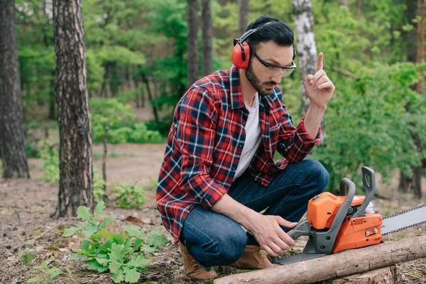 Holzfäller mit lärmintensiven Kopfhörern sitzt im Wald in der Nähe der Kettensäge und zeigt Ideengeste — Stockfoto