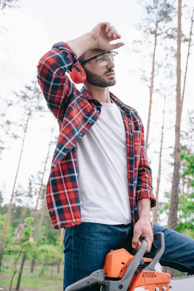 Lenhador cansado em óculos de proteção e auriculares segurando motosserra e olhando para longe na floresta — Fotografia de Stock