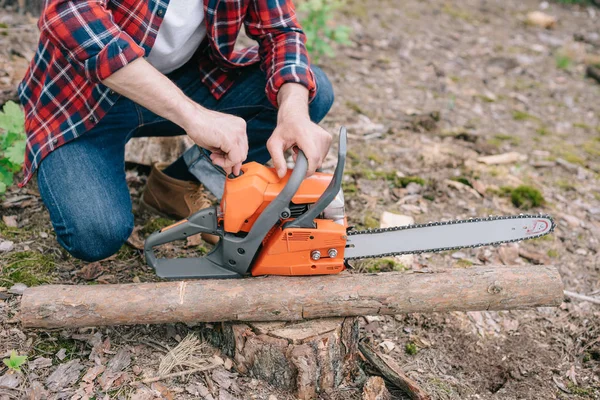 Teilansicht des Holzfällers, der die Kettensäge einstellt, während er in der Nähe des Baumstammes im Wald sitzt — Stockfoto