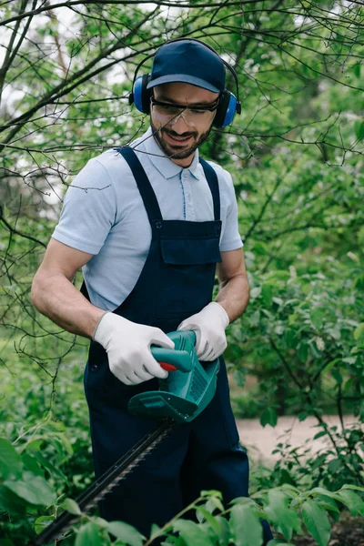 Jardinero sonriente en overoles y orejeras cortando arbustos con trimmer eléctrico en el parque - foto de stock
