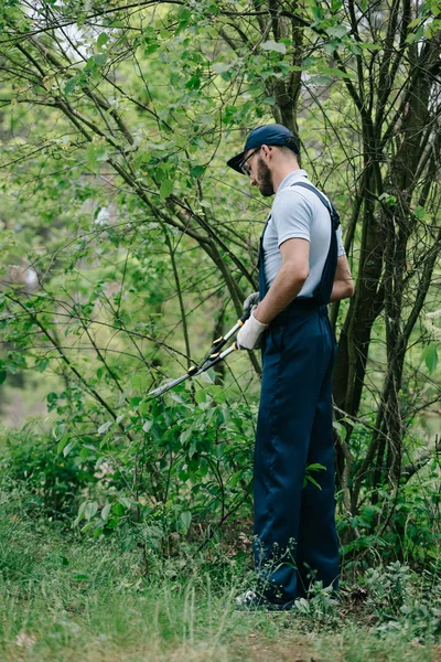 Gärtner in Overalls und Mütze Schneiden von Sträuchern mit Trimmer im Garten — Stockfoto