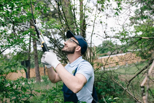 Jardinier souriant dans des lunettes de protection et des buissons de coupe de casquette avec tondeuse dans le parc — Photo de stock