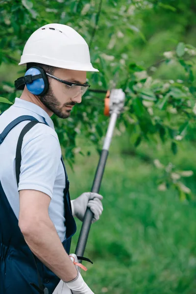 Selective focus of thoughtful gardener in helmet and hearing protectors trimming trees with telescopic pole saw in garden — Stock Photo