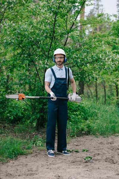 Handsome gardener in overalls, helmet and earmuffs holding telescopic pole saw and looking at camera — Stock Photo