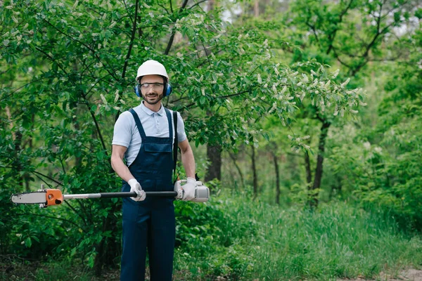 Jardinero alegre en overoles, casco y protectores auditivos sosteniendo la sierra telescópica y sonriendo a la cámara - foto de stock