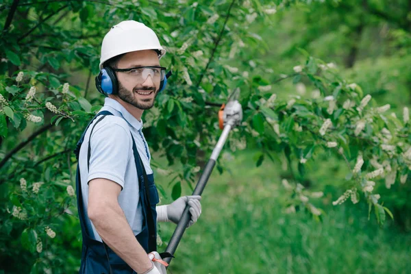 Hermoso jardinero en casco, gafas protectoras y auriculares que cancelan el ruido sosteniendo la sierra telescópica y sonriendo a la cámara - foto de stock