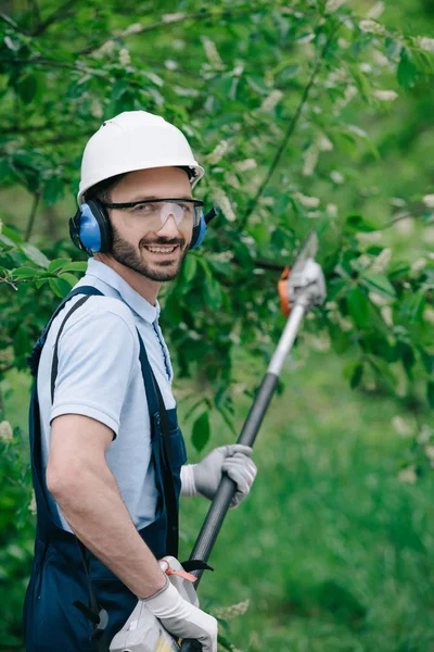 Jardinero alegre en casco, gafas protectoras y auriculares que cancelan el ruido sosteniendo la sierra telescópica y sonriendo a la cámara - foto de stock