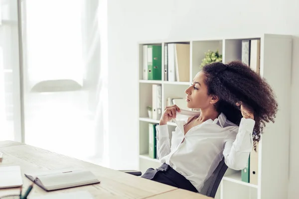 Bella donna d'affari afroamericana guardando altrove mentre seduto sul posto di lavoro e soffre di calore in ufficio — Foto stock