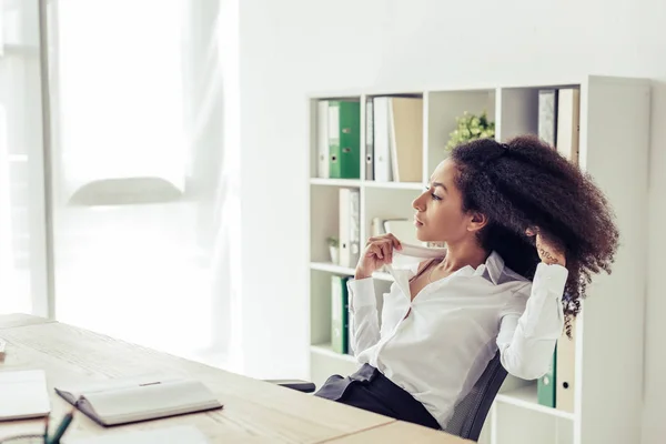 Bonita mujer de negocios afroamericana mirando hacia otro lado mientras está sentada en el lugar de trabajo y sufriendo de calor en la oficina - foto de stock