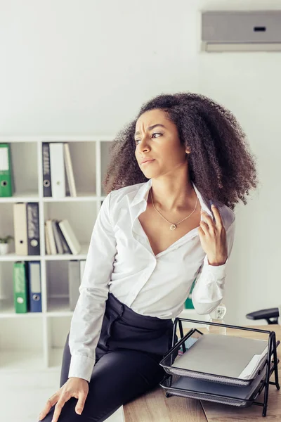 Erschöpfte afrikanisch-amerikanische Geschäftsfrau sitzt auf Schreibtisch, während sie im Büro unter Hitze leidet — Stockfoto