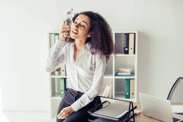 Pretty african american businesswoman suffering from heat and holding plastic bottle with water near head — Stock Photo