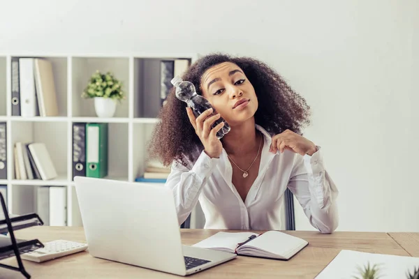 Pretty african american businesswoman looking at camera while sitting at workplace and holding plastic bottle near head — Stock Photo